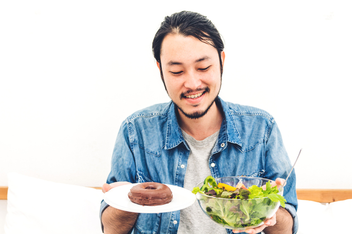 Man holding and making choice between healthy salad and calorie bomb chocolate donut on bed at home.Healthy eating and Junk food concept