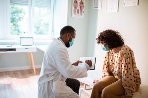 African American male doctor with surgical mask goes over paperwork in exam room with female Latina patient wearing surgical mask