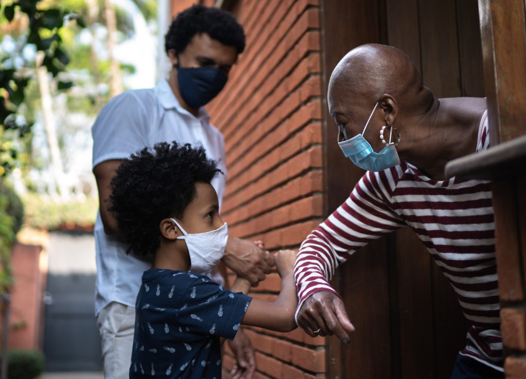 Black son and grandson wearing surgical masks greeting grandmother in surgical mask at door