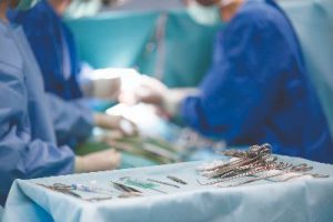 Gowned and gloved surgery staff with masks on prepare inside the operating room with surgical tools on a tray in the foreground