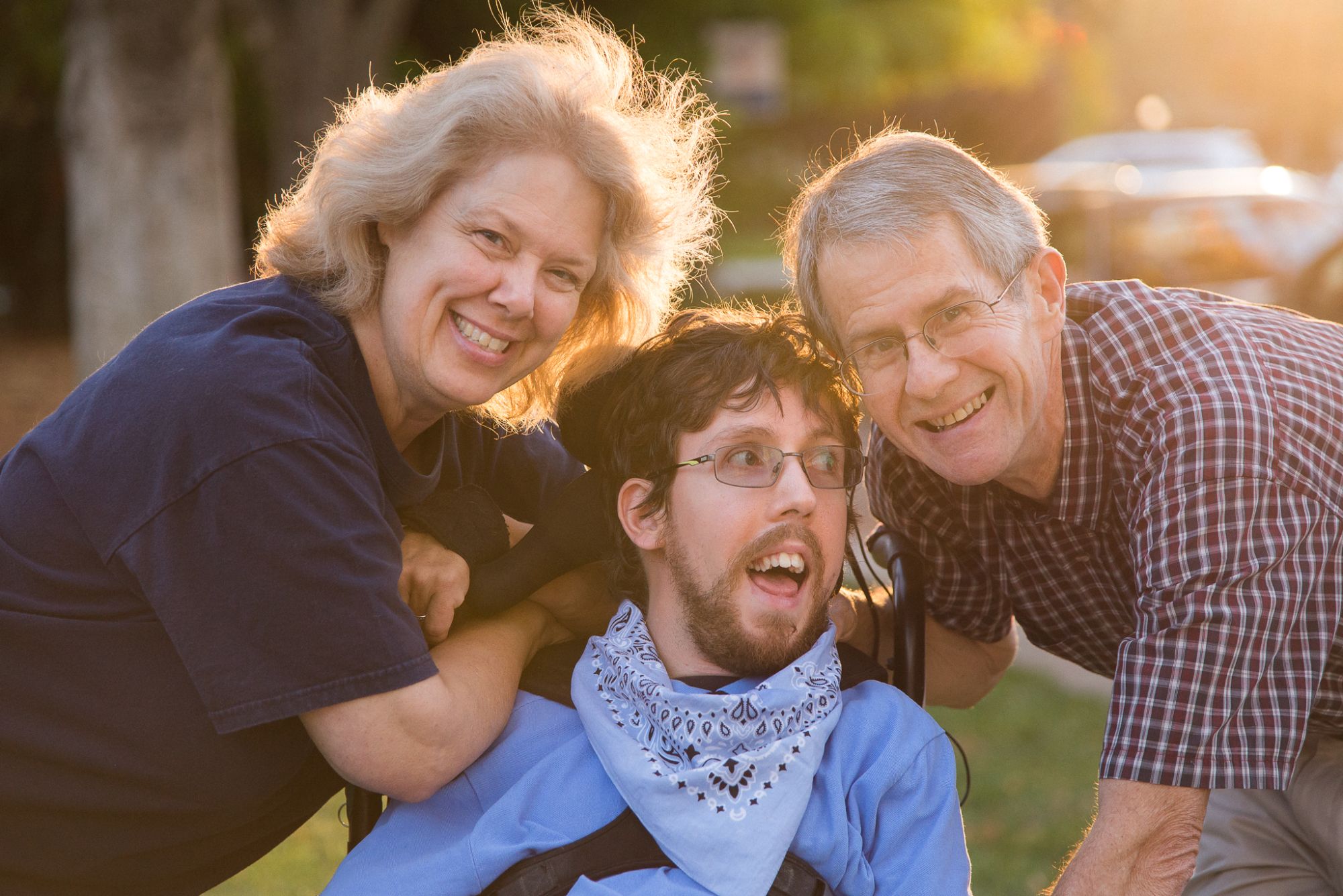 Parents pose with their son who is in a wheelchair on a sidewalk with the sun glowing behind them