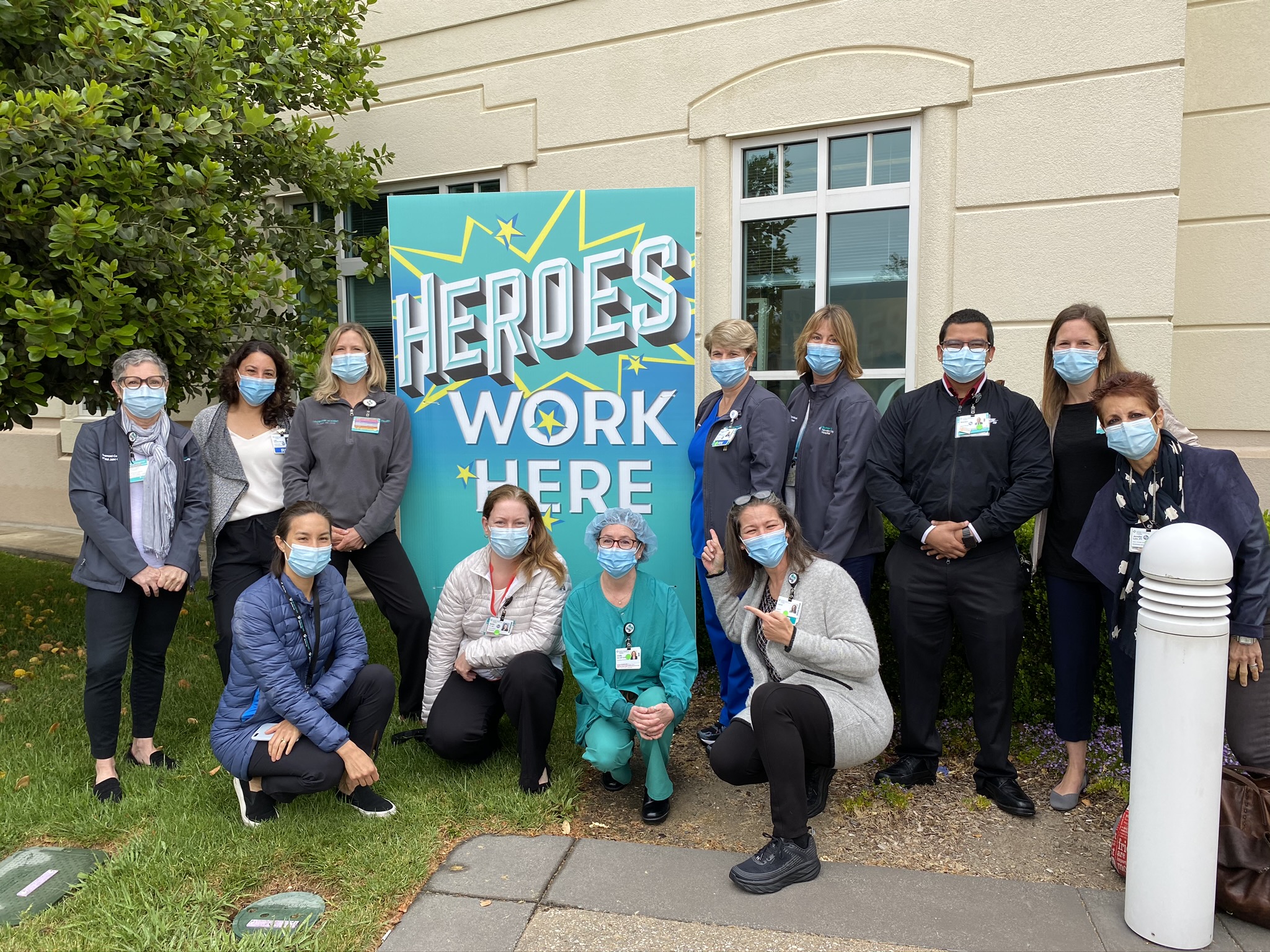 Medical staff at Novato Community Hospital are gathered around a 'Heroes Work Here' sign at the front of the hospital