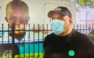 African American male with surgical mask stands alongside black iron fence and mobile healthcare van with African American boy's image