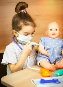 Photo of a child in a mask pretending to give a doll a shot in the arm.