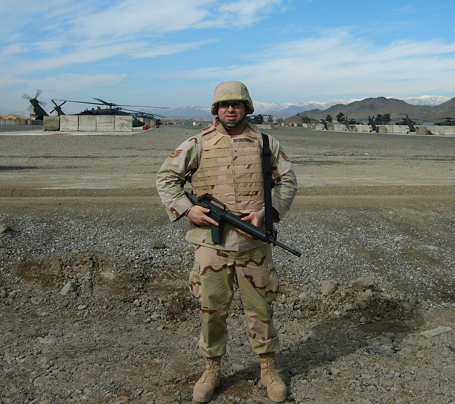 Man in army fatigues poses with rifle and military helicopter and mountains in the background