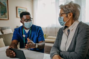 Black male nurse consults Latina patient while wearing surgical masks and gesturing to smart tablet