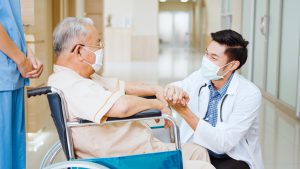Young male Asian doctor crouch down holding hand and talk to the senior adult patient on wheelchair in hospital hallway. Medical healthcare job, or hospital business concept
