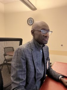 African American man with glasses sits in office looking at computer with wall clock behind him
