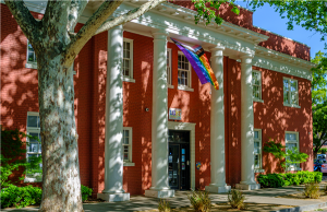 Facade of brick building exterior with triangular pediment and doric columns with LGBTQ+ rainbow pride flag draped outside