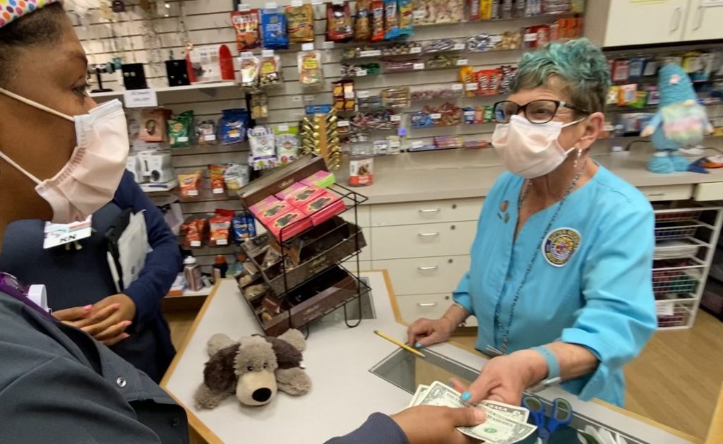 Volunteer Alyce Glazer, wearing a teal top and glasses and a white mask, helps black female gift shop customer make a purchase.