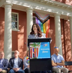 Woman with long brown hair stands at a podium in front of a red brick building with doric white columns and a multicolored flag behind her.