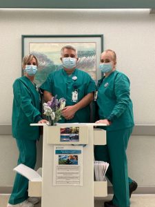 Registered nurses in teal scrubs and surgical masks gather around rolling utility cart