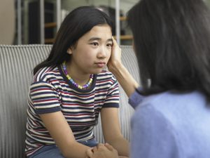 Asian adolescent girl with striped top and multicolored necklace talking with concerned mother on couch