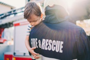 Firefighter holds young boy with striped shirt who has closed eyes and hangs over firefighters shoulder stock photo