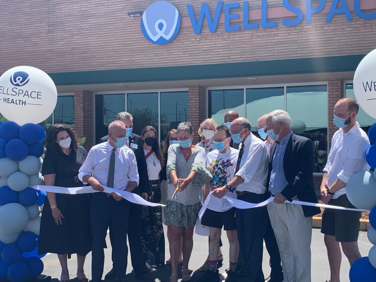 Multiple adults wearing surgical masks standing in front of a brick health clinic cutting a ceremonial while ribbon