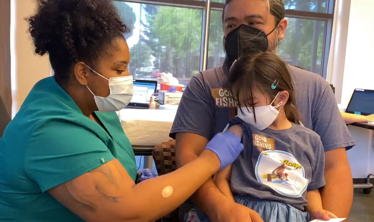 A Sutter health medical professional gives a COVID vaccination to a little girl who is sitting on her father's lap. They are both wearing matching "Gone Fishing Curious George" t-shirts.