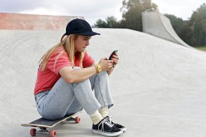 Caucasian female with black baseball cap red t-shirt and-jeans sits on skateboard looking at her phone at a skate park stock-photo