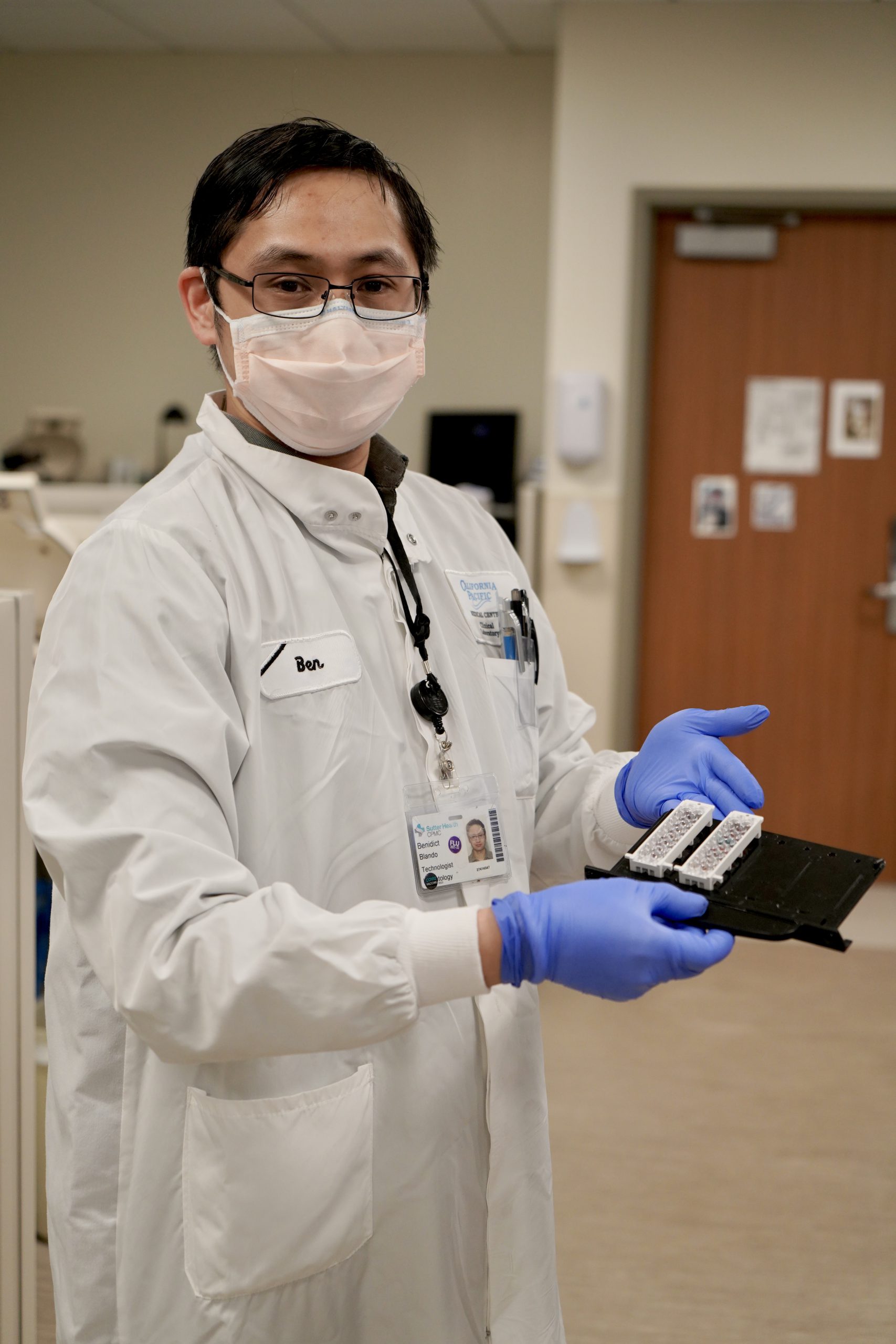 A young Asian scientist in a white lab coat, blue medical gloves and a mask, holds blood testing equipment inside a hospital blood bank laboratory. 