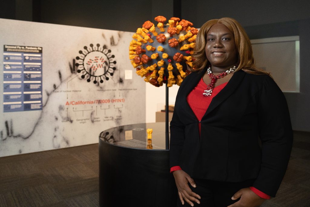 Black woman with black suit and red blouse stands in front of a 3D coronavirus model