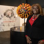 Black woman with black suit and red blouse stands in front of a 3D coronavirus model