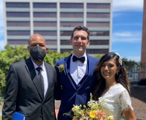 Couple get married with officiant. Officiant in suit, groom in tux, bride in wedding dress holding flowers.