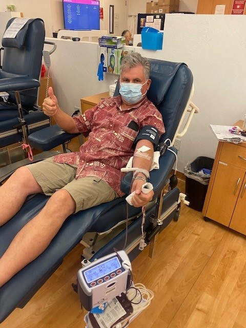 Mark Lindberg, a white male in a Hawaiian shirt and cargo shorts sits is a chair while making a blood donation. 