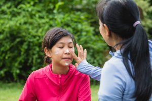 Asian mother applying sunscreen lotion to protect her daughter's face before exercise while setting on floor in the park at outdoors - stock photo