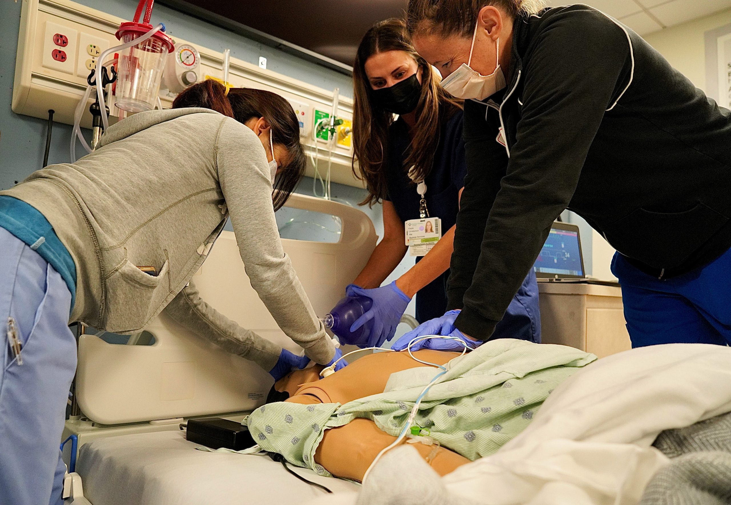 Three nurses are gathered around a mock patient bed as they practice administering air on a simulation mannequin. 