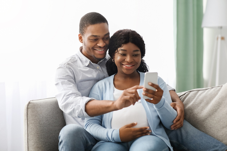 Loving black expecting parents using mobile application on smartphone, reclining together on couch and embracing, looking at mobile phone screen and smiling