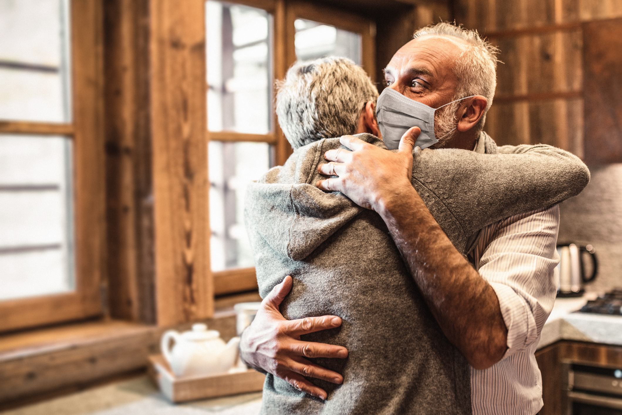 Older Caucasian couple embracing in a cabin kitchen with white-haired man with surgical mask looks to the window