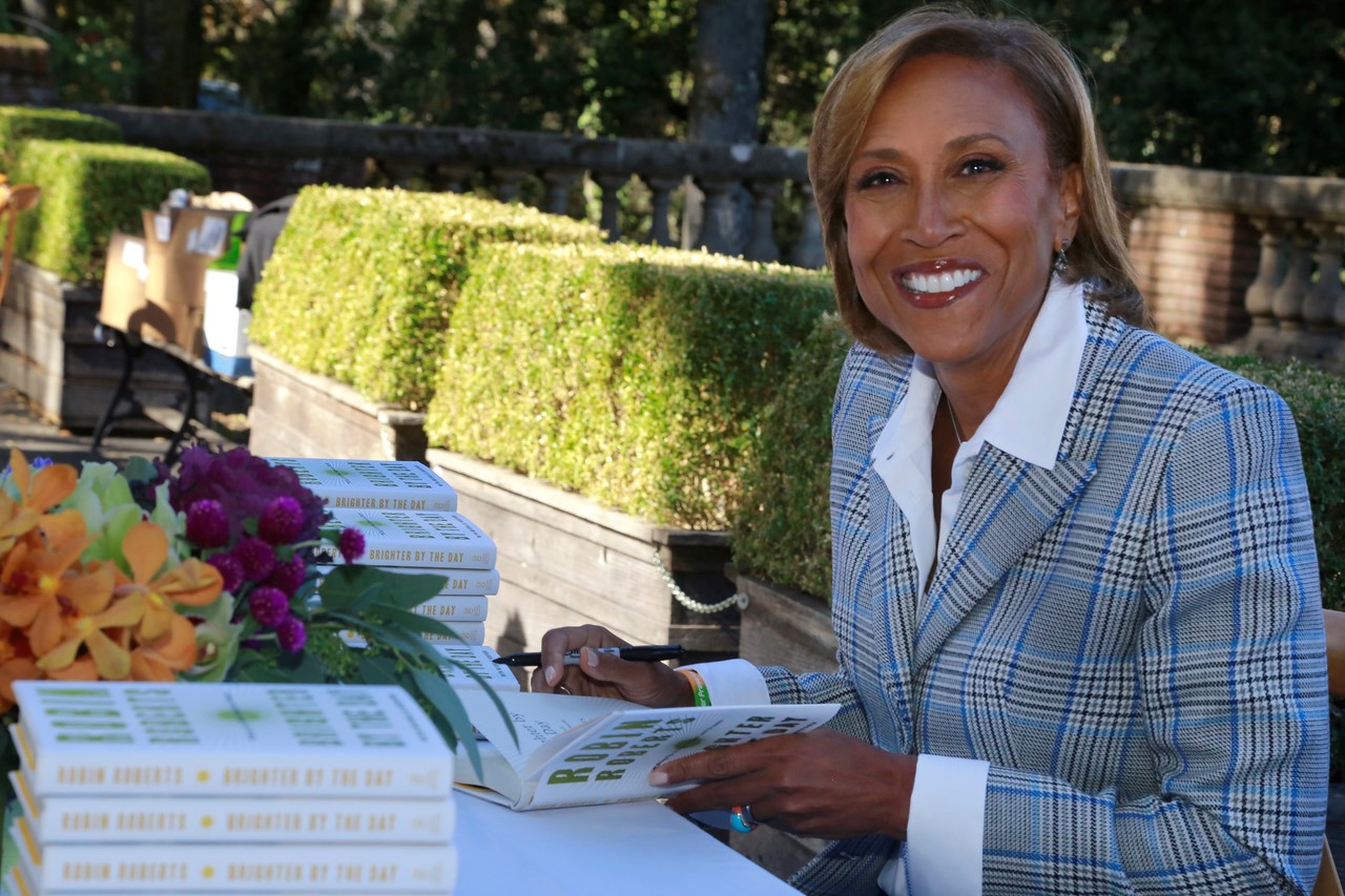 Robin Roberts signing books in striped suit with white blouse smiles at the camera while she signs books on a table with a white tablecloth and flower arrangement in the corner.