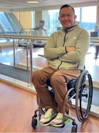 Steve Lau, an Asian American man, sits in a wheelchair with his arm folded and a big smile inside Sutter's Alta Bates Summit Medical Center acute rehab services.