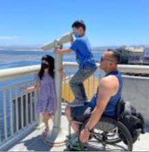 Steve Lau in a wheelchair with his two young children, a young son and daughter, who are outside on a p
