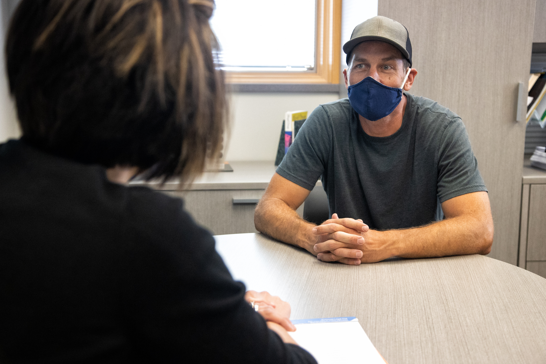 A Female Mental Health Professional Talks with a Masked White Male in His Forties while Sitting at a Table in a Medical Clinic