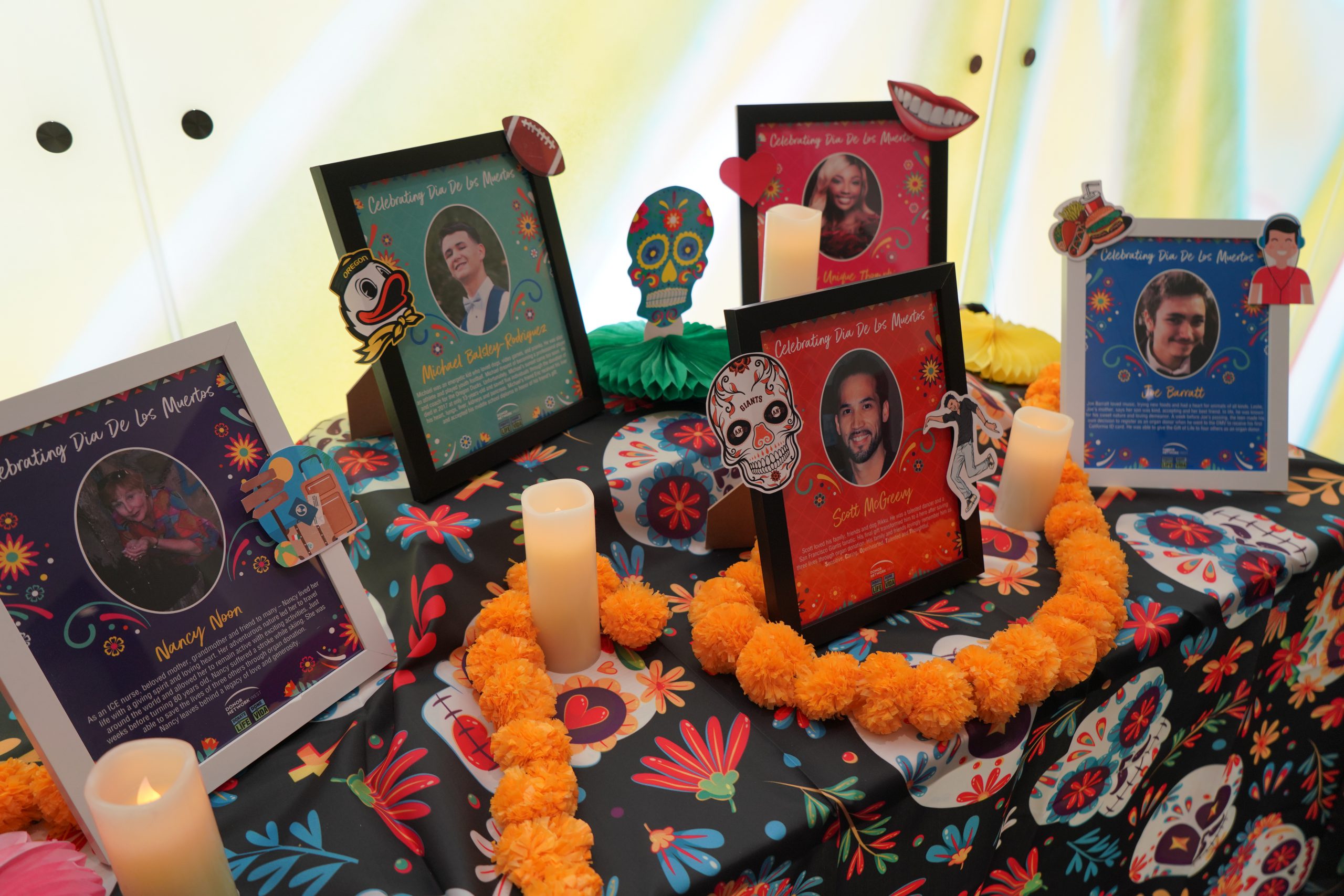 A traditional Day of the Dead altar or ofrendas is adorned with framed stories of individuals whose organs were used to give the gift of life to someone in need. The table has a colorful Day of the Dead-themed tablecloth, battery powered candles and faux marigold flowers. 