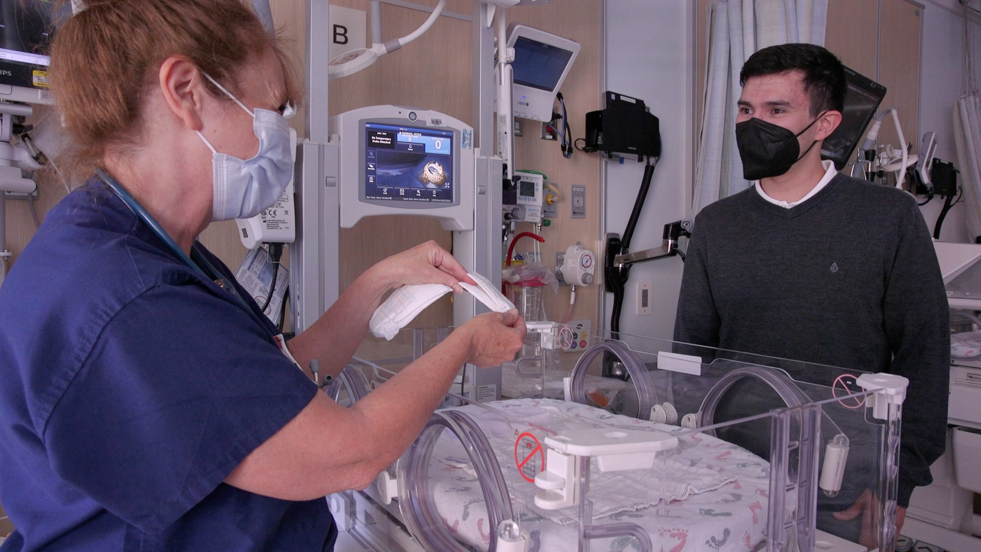 Zach and nurse stand in a NICU room next to an isolette designed to keep preemie babies warm. 