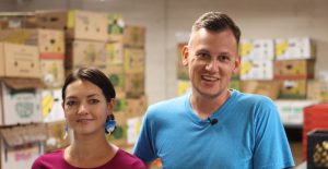 Caucasian woman with brown hair tied back and red shirt and a Caucasian man with short blond hair and blue shirt stand in front of numerous grocery produce boxes