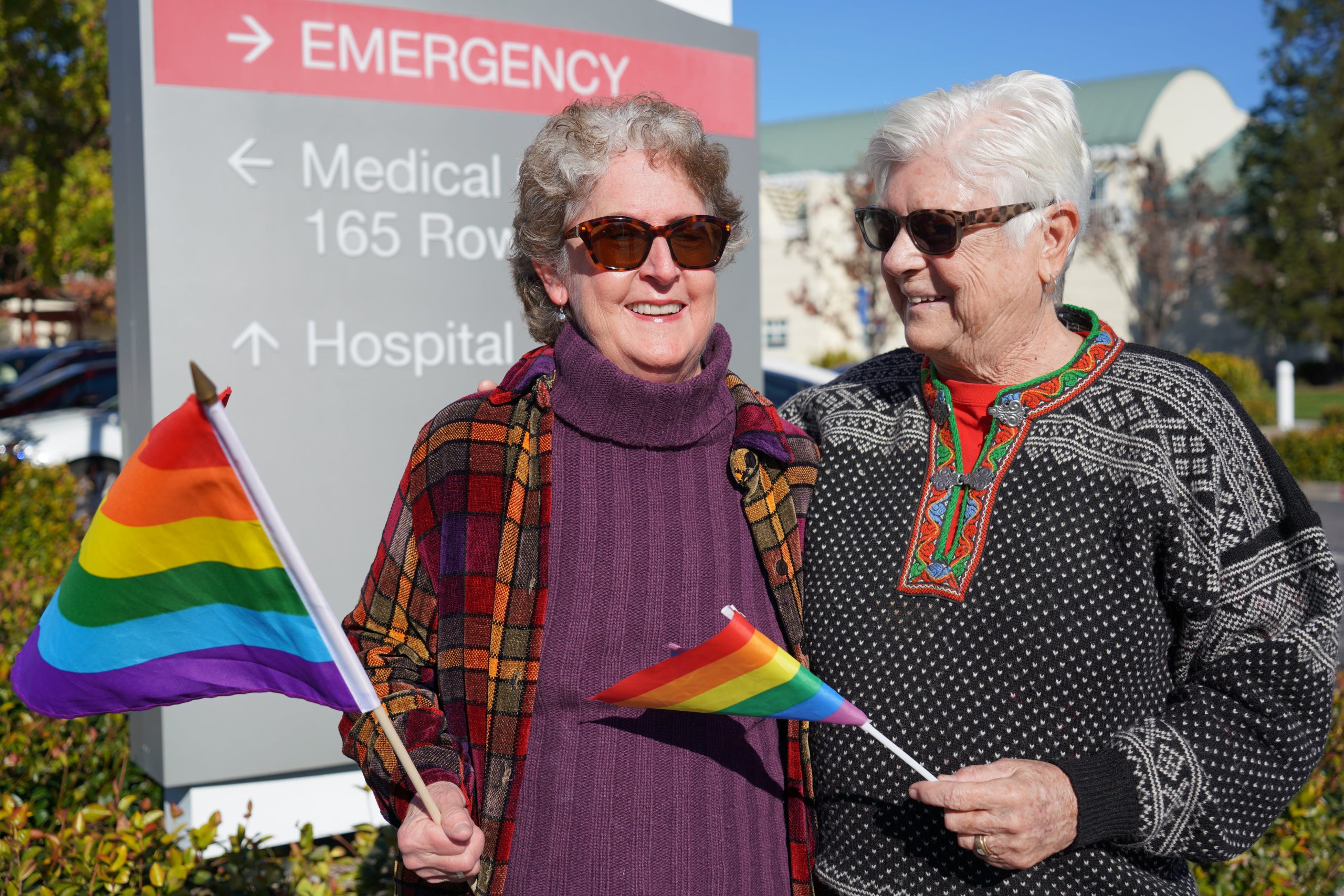 Lolma Olson (pictured left) and Beth Reed (pictured right) stand in front of the hospital emergency entrance sign while holding rainbow flags in support of the LGBTQ community.