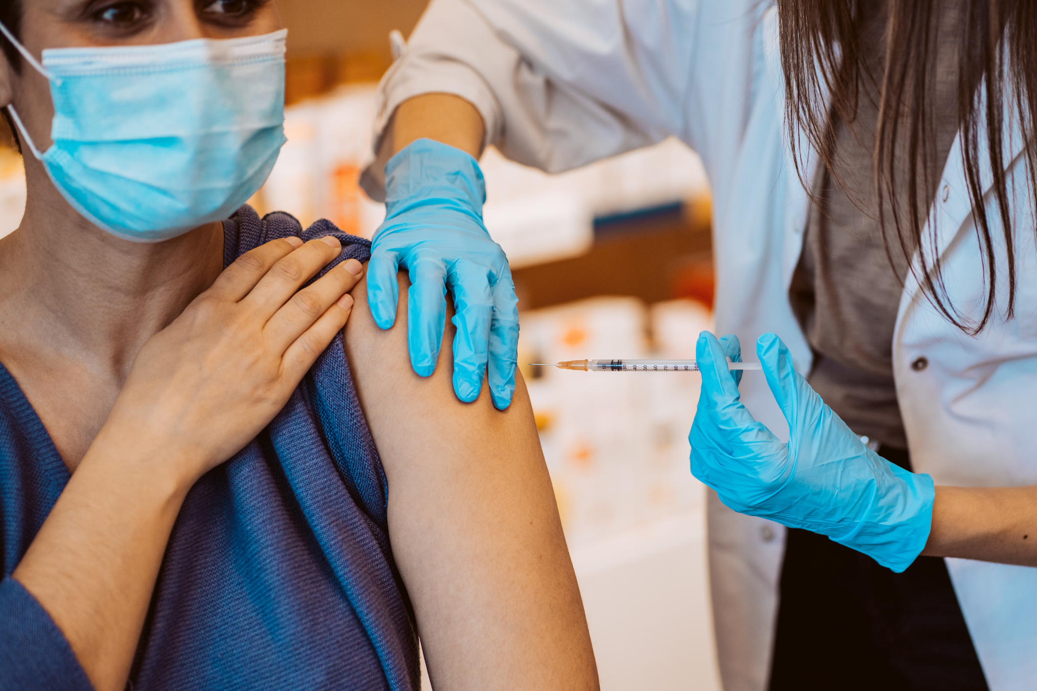 Latina woman wearing surgical mask receives vaccination shot from long brown haired woman wearing white lab coat and blue surgical gloves