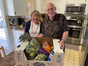 An older Caucasian woman and man posing in kitchen with a box of fresh fruits and vegetables in front of them,