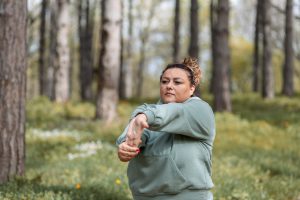 Middle-aged, plus-sized Latina woman stretching her arm before her morning run in a wooded area on a path. 