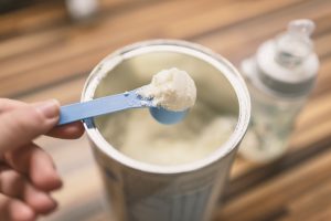 Powdered infant formula scooped out of canister with blue scoop on wooden countertop with baby bottle in the background.