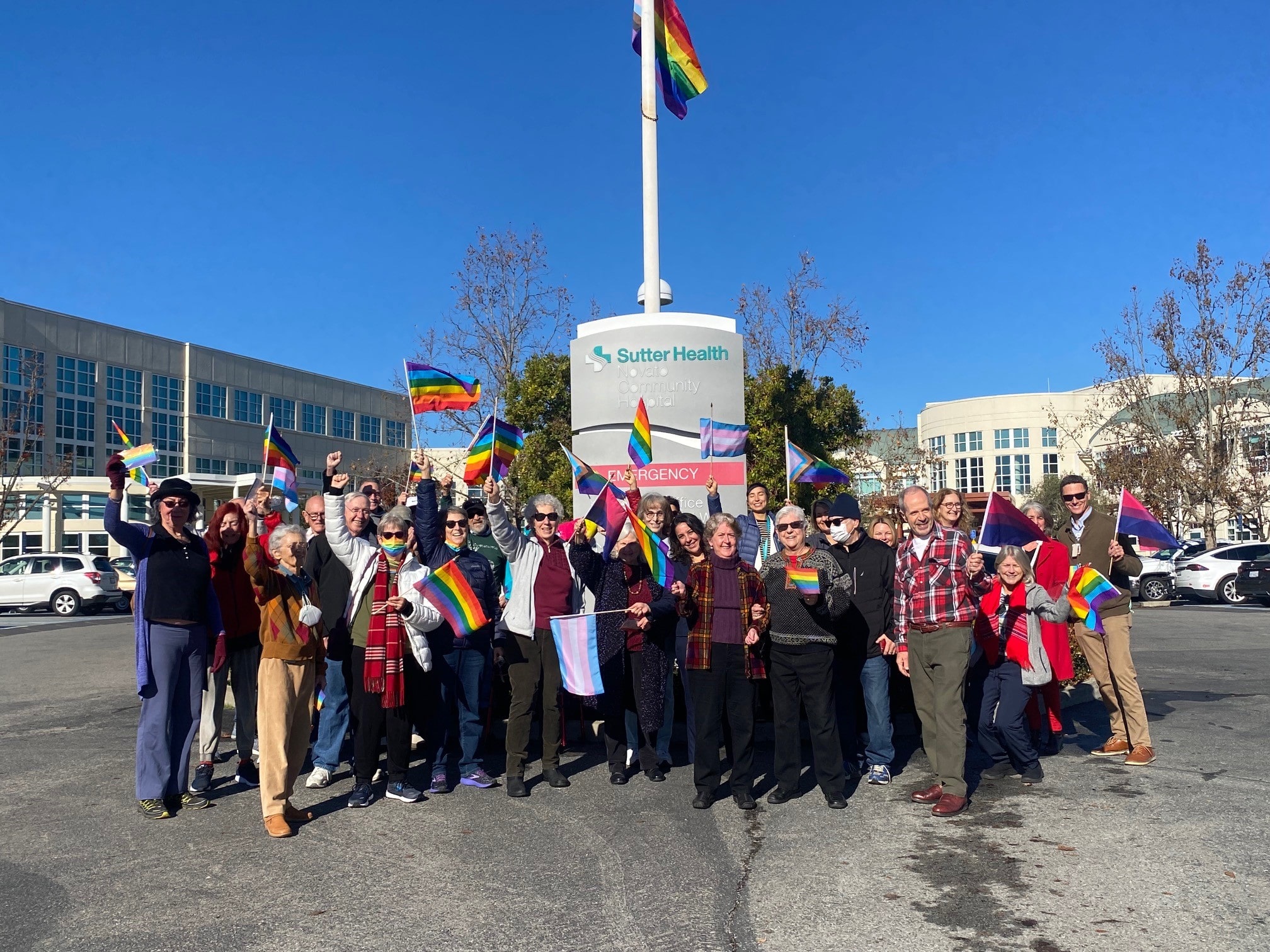 A group of about 30 individuals hold rainbow-colored pride flags outside of Novato Community Hospital. It is a sunny day. 