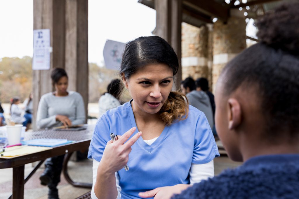 Black adult mother uses digital tablet at a table in the background while a Latina female doctor in light blue scrubs gestures and speaks with a young Black girl at a picnic table at an outdoor community health clinic.