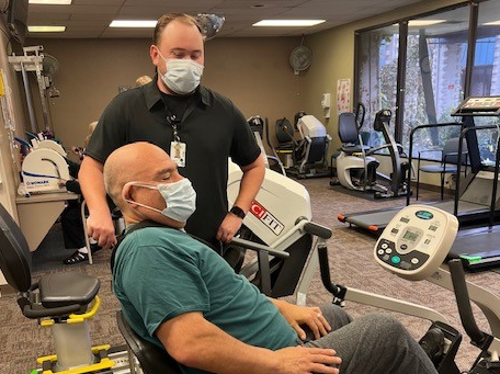 Caucasian male with surgical mask and black button-down shirt stands over bald Latino man with green t-shirt on seated on an exercise bike.