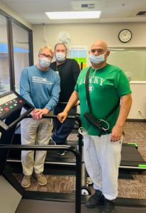 Three men with surgical masks stand on or around a treadmill facing the camera
