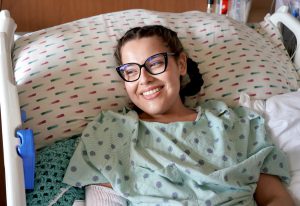 A Hispanic woman in her late 20s sits up in a hospital bed wearing a hospital gown. She is smiling and is wearing black glasses. Her hair is in two braids.