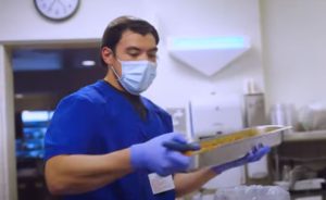 Man in blue scrubs with surgical mask and gloves handles a tray of food in a hospital kitchen.