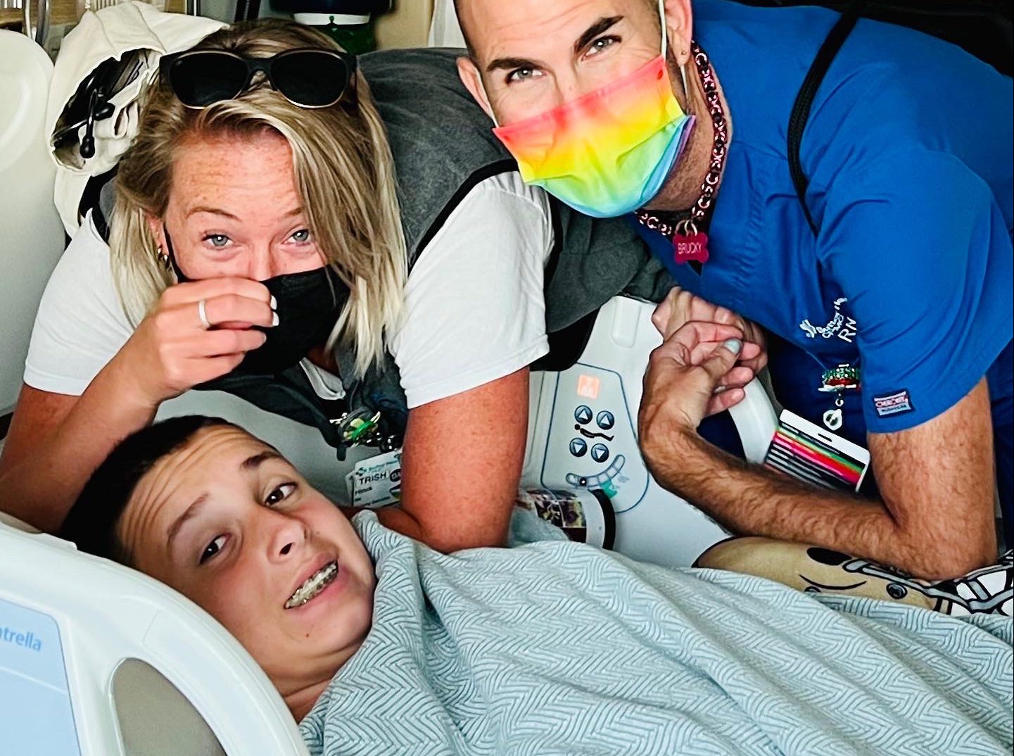 Teenage boy in a hospital bed smiles with is mom and nurse in a tie-dye mask next to him.