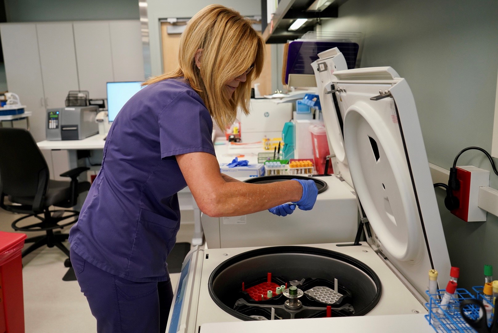 A Caucasian woman in purple scrubs removes a blood sample from a large cyclotron in a hospital laboratory.